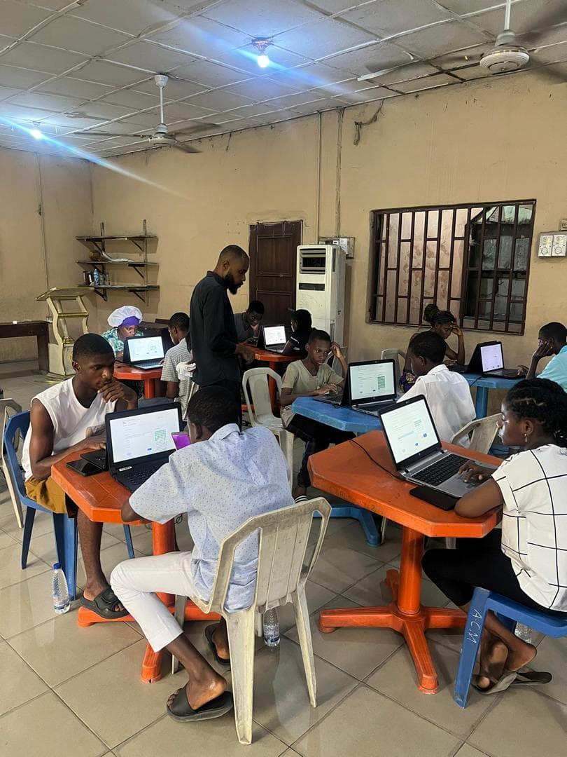 Students sit at laptops working through computer-based JAMB practice questions.
